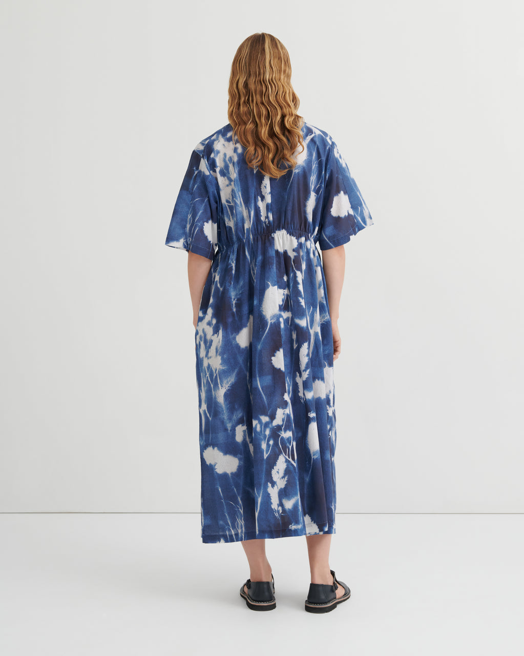 A woman viewed from behind wearing a loose, short-sleeve midi dress with a blue and white tie-dye pattern, paired with black flat sandals, against a plain white studio background.