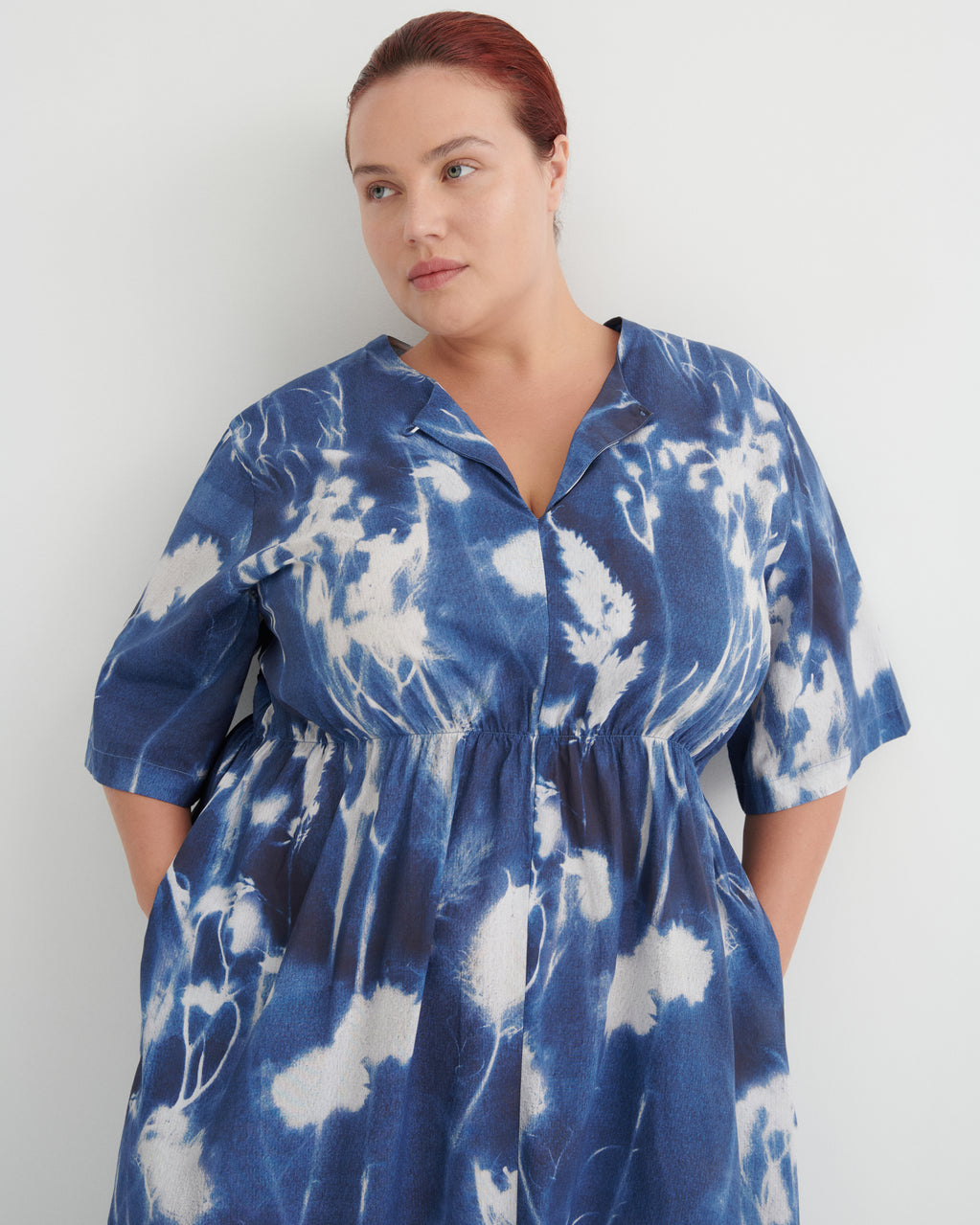 A woman wearing a blue and white tie-dye short-sleeve shirt dress, standing against a plain light gray wall.