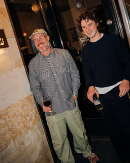 Two men standing in a doorway at night, holding wine glasses. The man on the left wears a beige cap, a gray and white vertically striped button-down shirt, and olive green cargo pants.