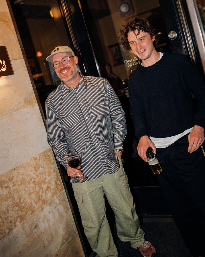Two men standing in a doorway at night, holding wine glasses. The man on the left wears a beige cap, a gray and white vertically striped button-down shirt, and olive green cargo pants.