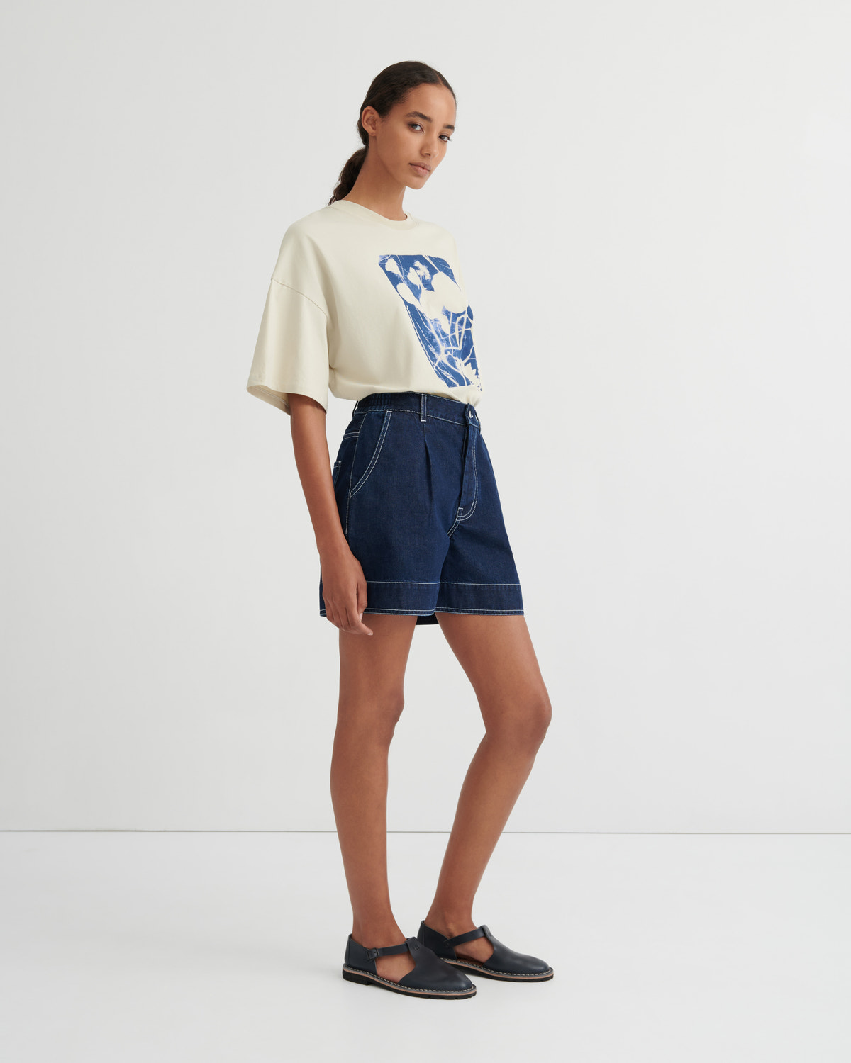 A model stands in a studio wearing a cream-colored oversized t-shirt with a blue graphic print, dark blue denim shorts, and dark navy Mary Jane flats.