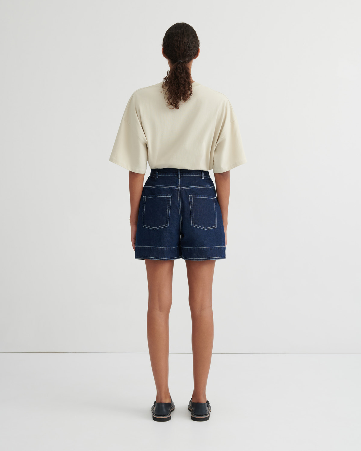 A model viewed from behind wearing a cream-colored short-sleeve crop top and dark blue denim shorts with contrast stitching, paired with dark loafers, against a plain white studio background.