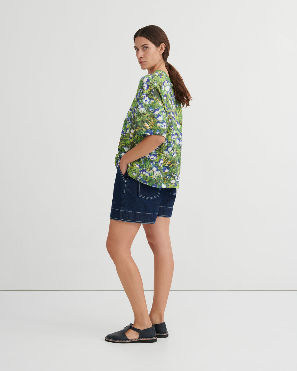 A woman stands in profile wearing a green short-sleeve button-up shirt with a blue and white floral print, dark denim shorts, and black flat sandals against a plain light gray studio background.