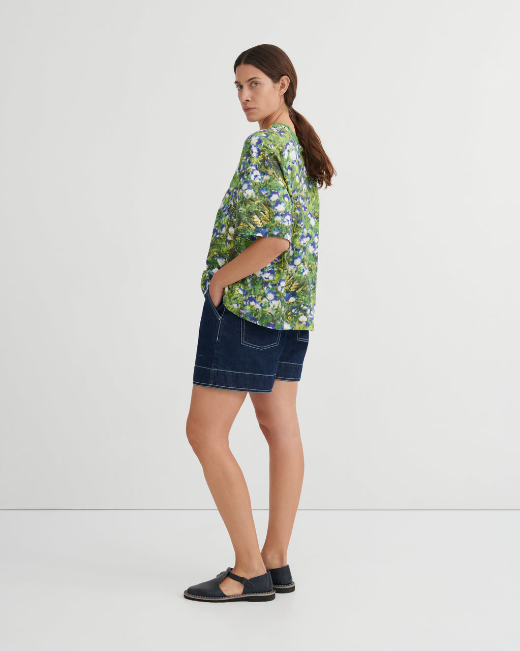 A woman stands in profile wearing a green short-sleeve button-up shirt with a blue and white floral print, dark denim shorts, and black flat sandals against a plain light gray studio background.