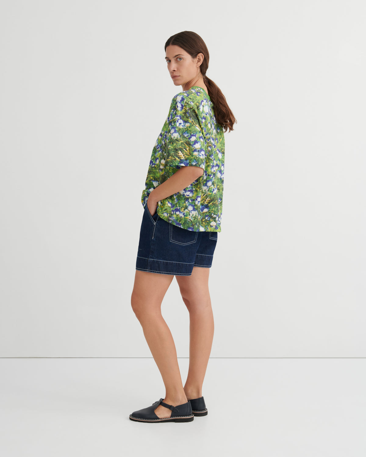 A woman stands in profile wearing a green short-sleeve button-up shirt with a blue and white floral print, dark denim shorts, and black flat sandals against a plain light gray studio background.