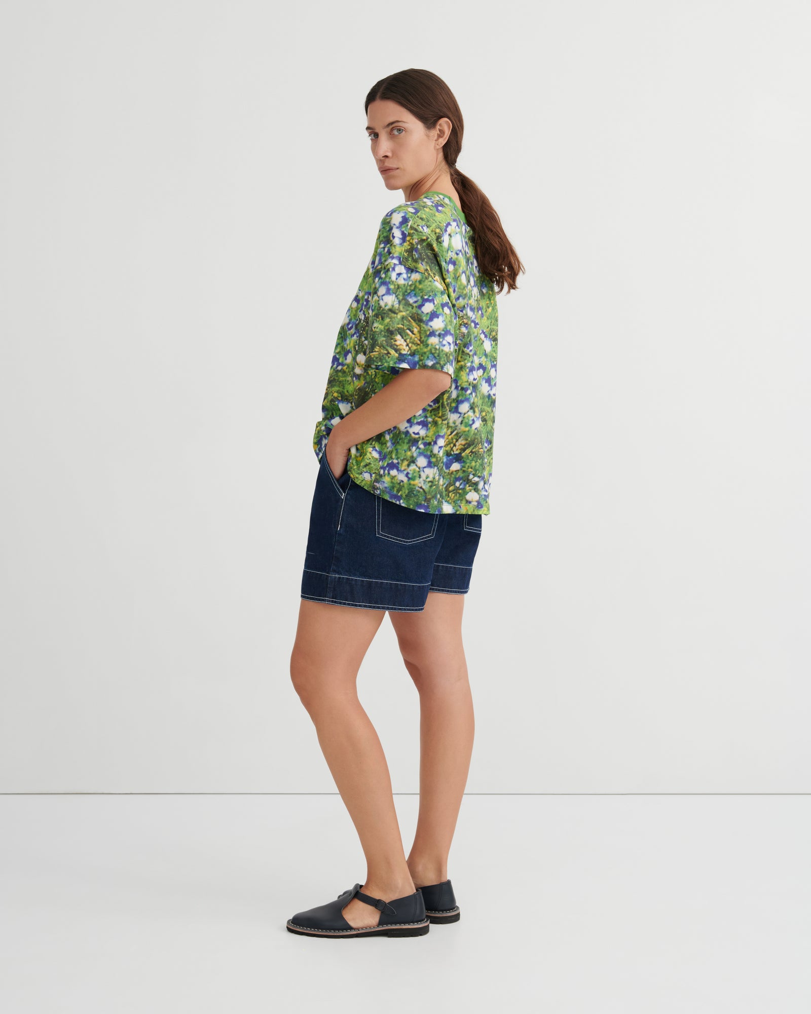 A woman stands in profile wearing a green short-sleeve button-up shirt with a blue and white floral print, dark denim shorts, and black flat sandals against a plain light gray studio background.
