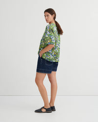 A woman stands in profile wearing a green short-sleeve button-up shirt with a blue and white floral print, dark denim shorts, and black flat sandals against a plain light gray studio background.