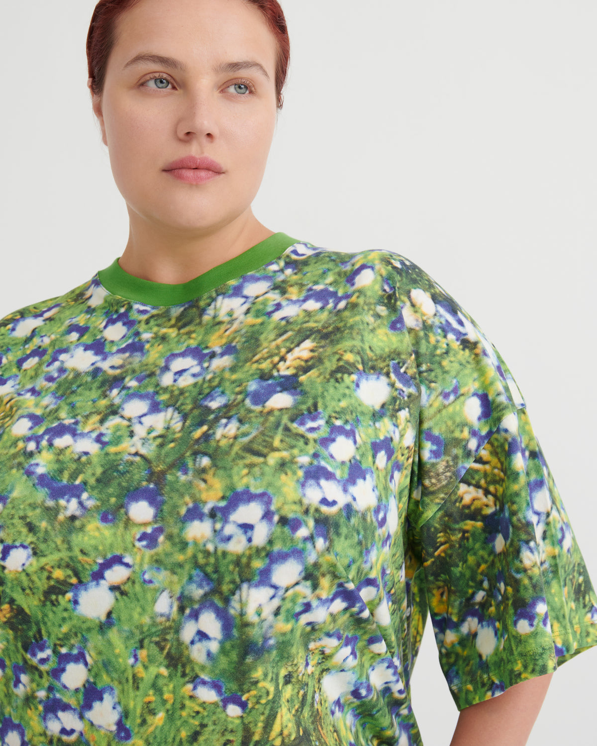 A close-up of a model wearing a short-sleeve crewneck t-shirt with an all-over floral print, featuring green, blue, and white blossoms on a textured green background.