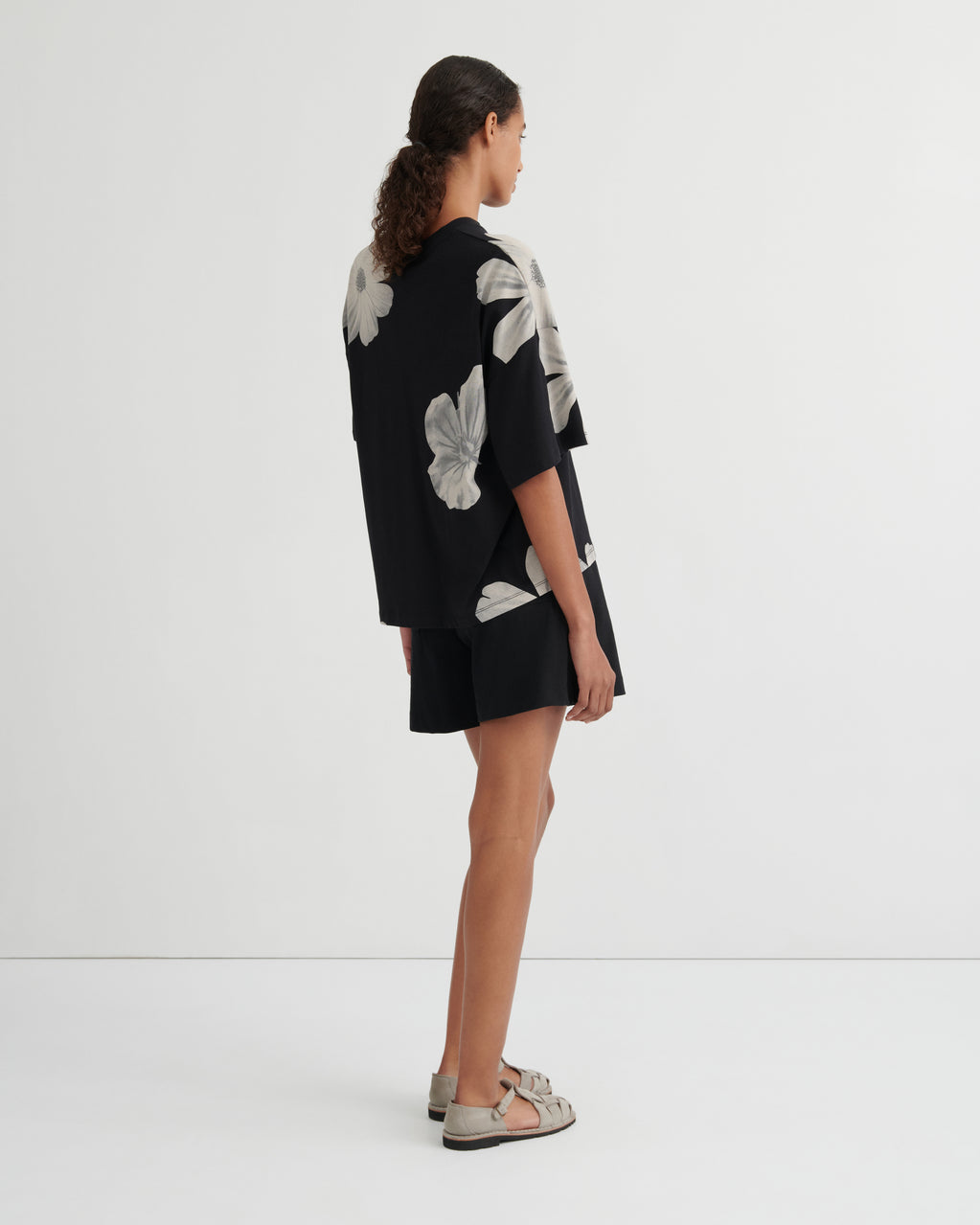 A woman shown from the back wearing a black short-sleeve tunic top with large light gray floral prints, paired with black shorts and beige flat sandals, against a plain white studio background.