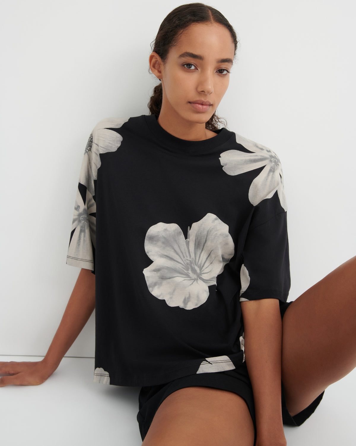 A woman wearing a black short-sleeve t-shirt with a large white hibiscus floral print, sitting against a plain white wall.