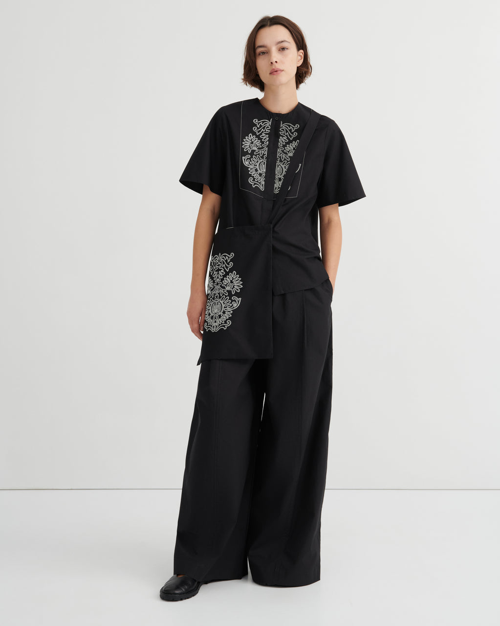 A model stands against a light gray studio background wearing a black short-sleeve top with white floral embroidery and matching black wide-leg pants.