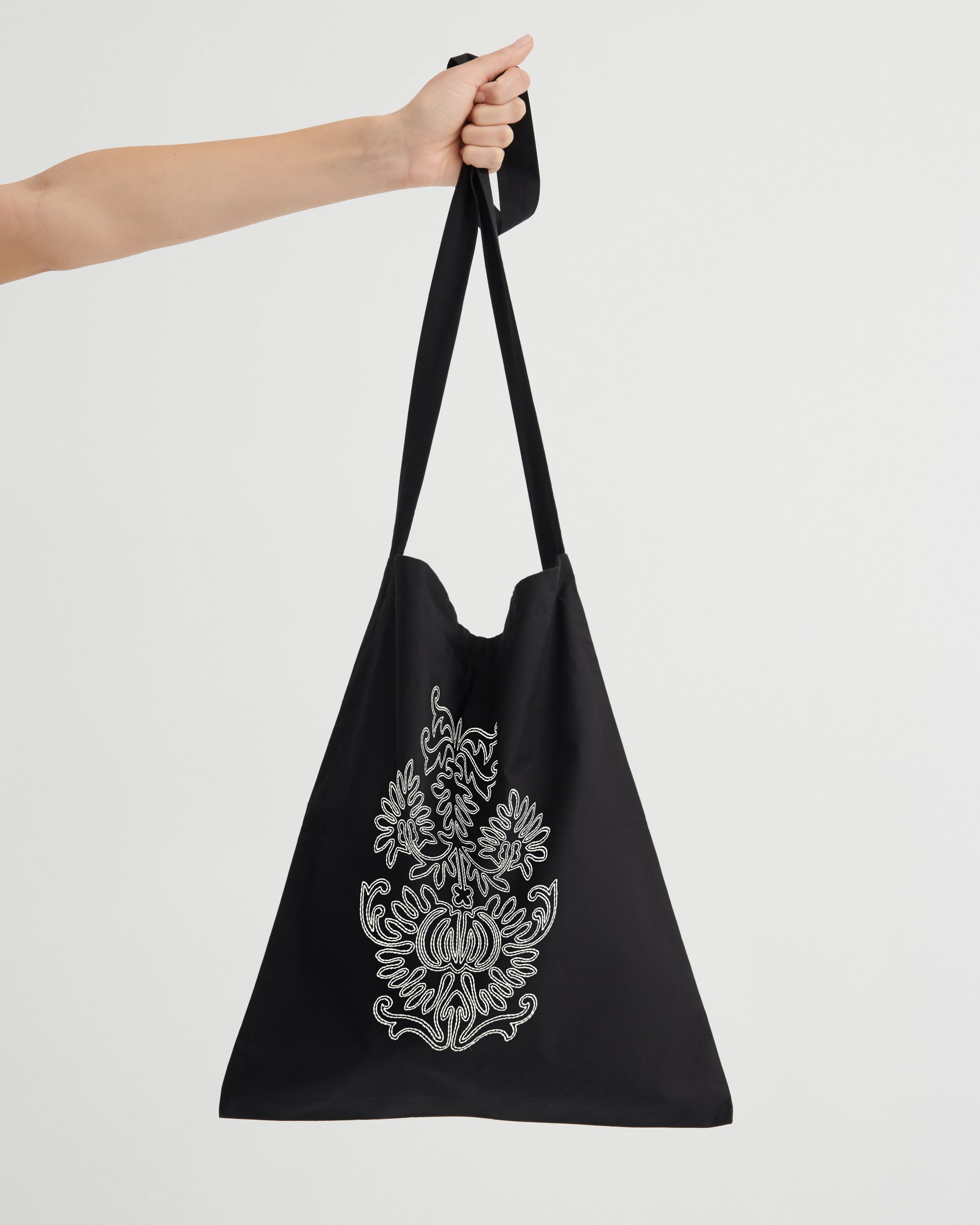 A black tote bag with a long shoulder strap, held by a person's arm against a light gray background. The bag features a white ornate floral graphic embroidered design.