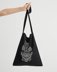 A black tote bag with a long shoulder strap, held by a person's arm against a light gray background. The bag features a white ornate floral graphic embroidered design.
