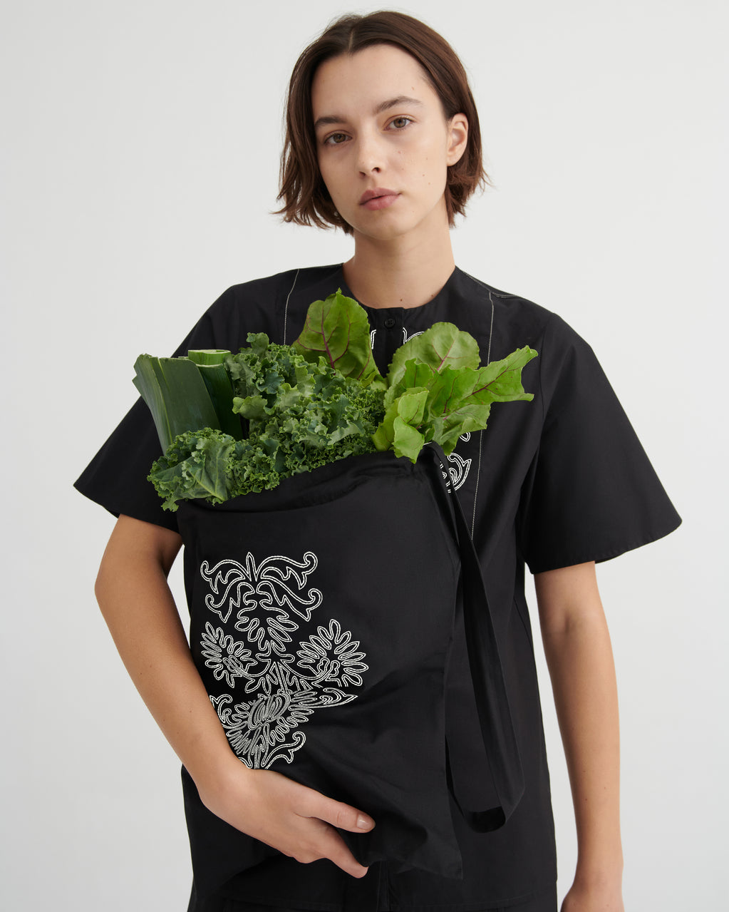 A model wearing a black short-sleeve t-shirt with white floral embroidery, holding a black fabric grocery bag filled with green kale and lettuce.