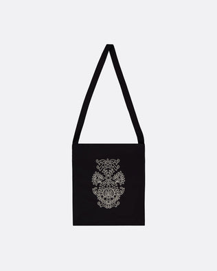 A black canvas shoulder bag with a long strap, featuring a white ornate damask-style pattern printed on the front, set against a plain light gray background.