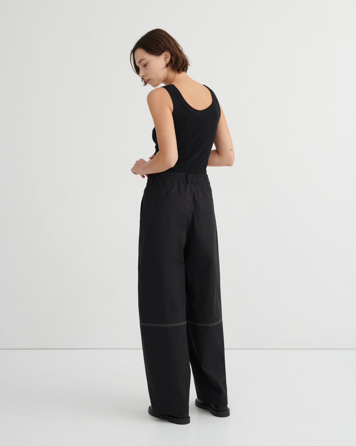 A woman wearing a black sleeveless top and black wide-leg trousers, photographed from the back against a clean white studio background.