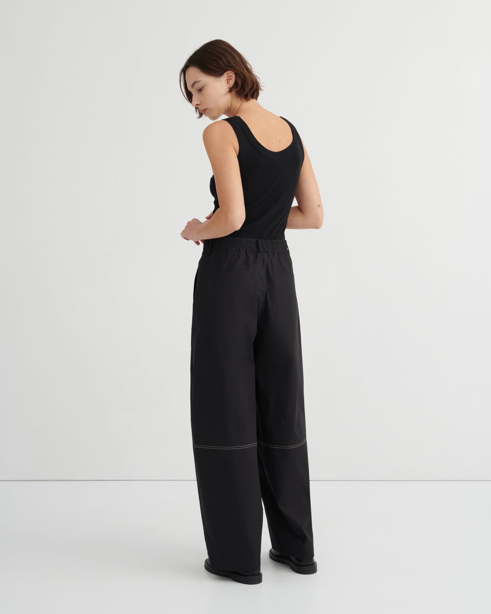 A woman wearing a black sleeveless top and black wide-leg trousers, photographed from the back against a clean white studio background.