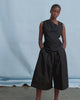 A women wears a black asymmetric top and wide leg black pant against a blue backdrop.