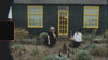 Super 8 camera footage of a black cottage with yellow trims and two people sitting outside it in the garden.