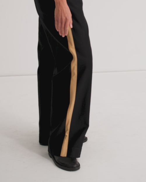A women wears a close fitting black scoop neck tank and relaxed black pants with a tan stripe down the side and black flats.