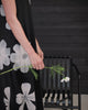 A model wearing a black sleeveless dress printed in white flowers sits on a black chair in front of black weatherboard backdrop.