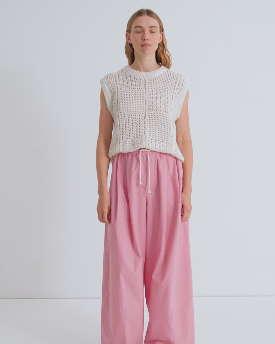A women wears a cream crochet detail vest and pink pleated trousers with a cream drawcord against a light grey studio background.
