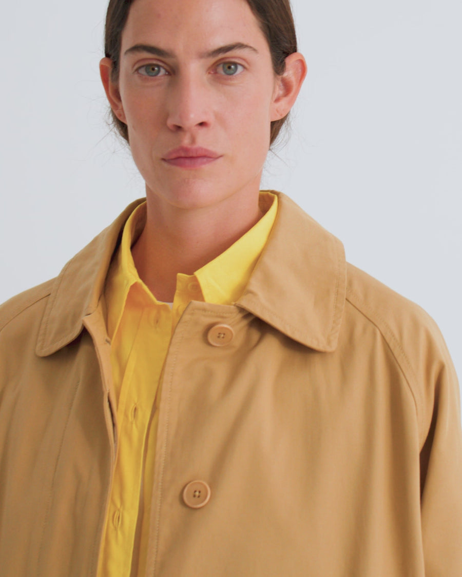 A women wears an unbuttoned, relaxed tan longline trench coat with a yellow shirt underneath and tan trousers.