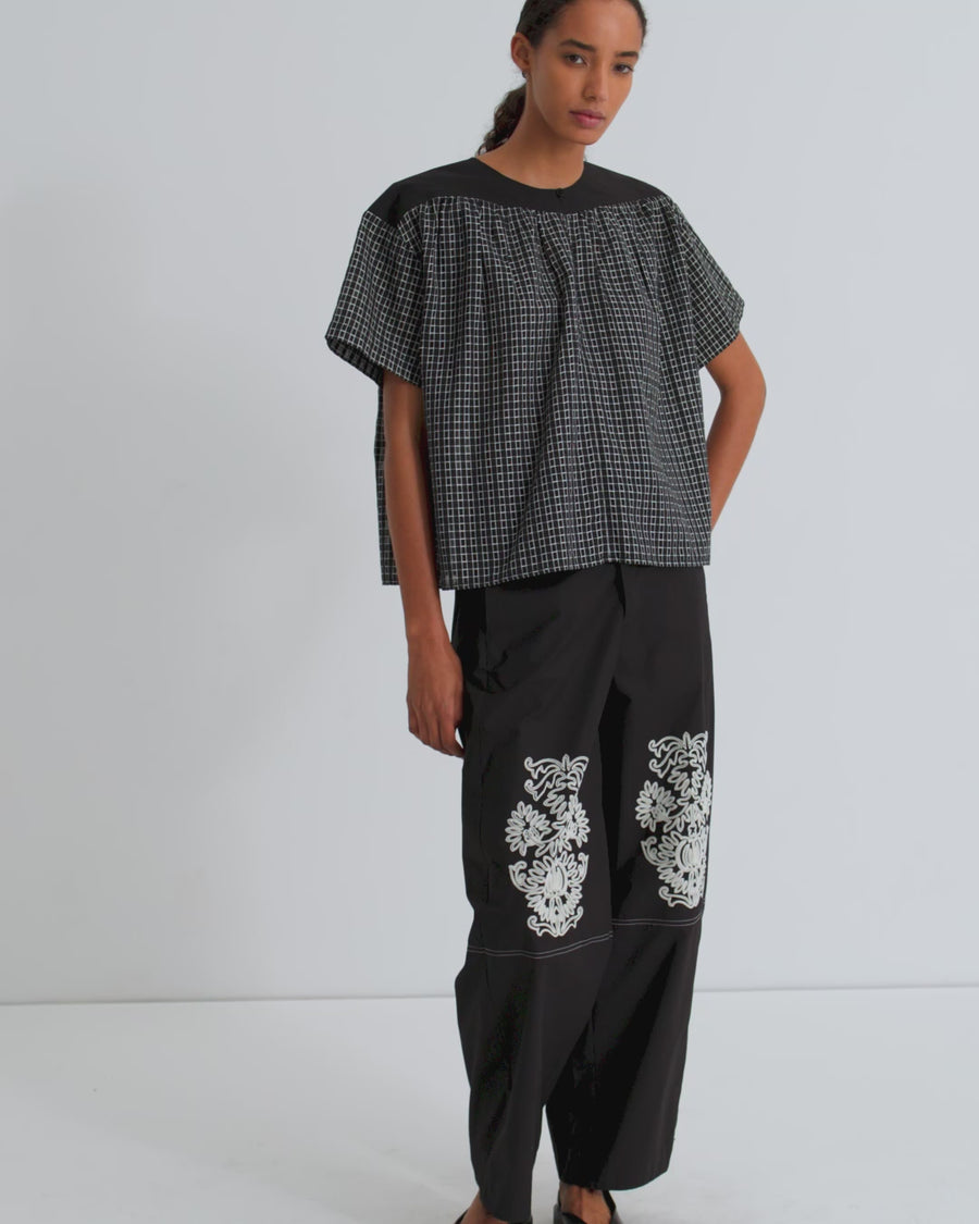A women wearing a black check top and black pants with floral embroidery on the  knee.