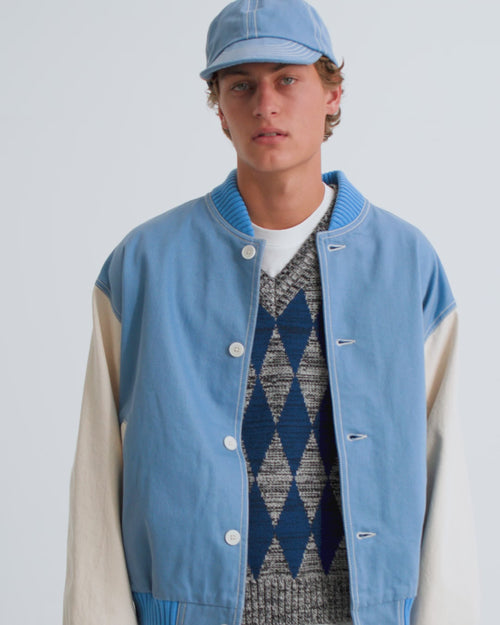 A man wears a blue cap, blue varsity jacket and patterned v neck sweater vest with dark blue jeans.