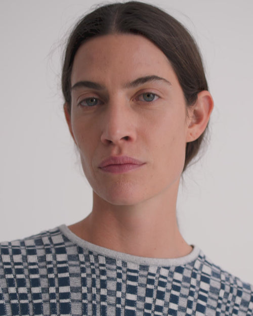 A women wearing a check blue and grey knitted top and dark denim shorts against a grey background.