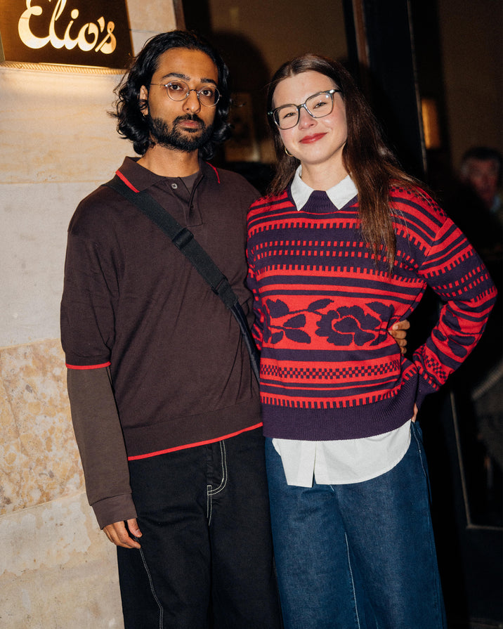Two people standing together in front of a building entrance at night. The person on the right wears a red and navy patterned sweater over a white collared shirt and blue jeans, while the person on the left wears a dark brown long-sleeve polo with red trim and black pants.