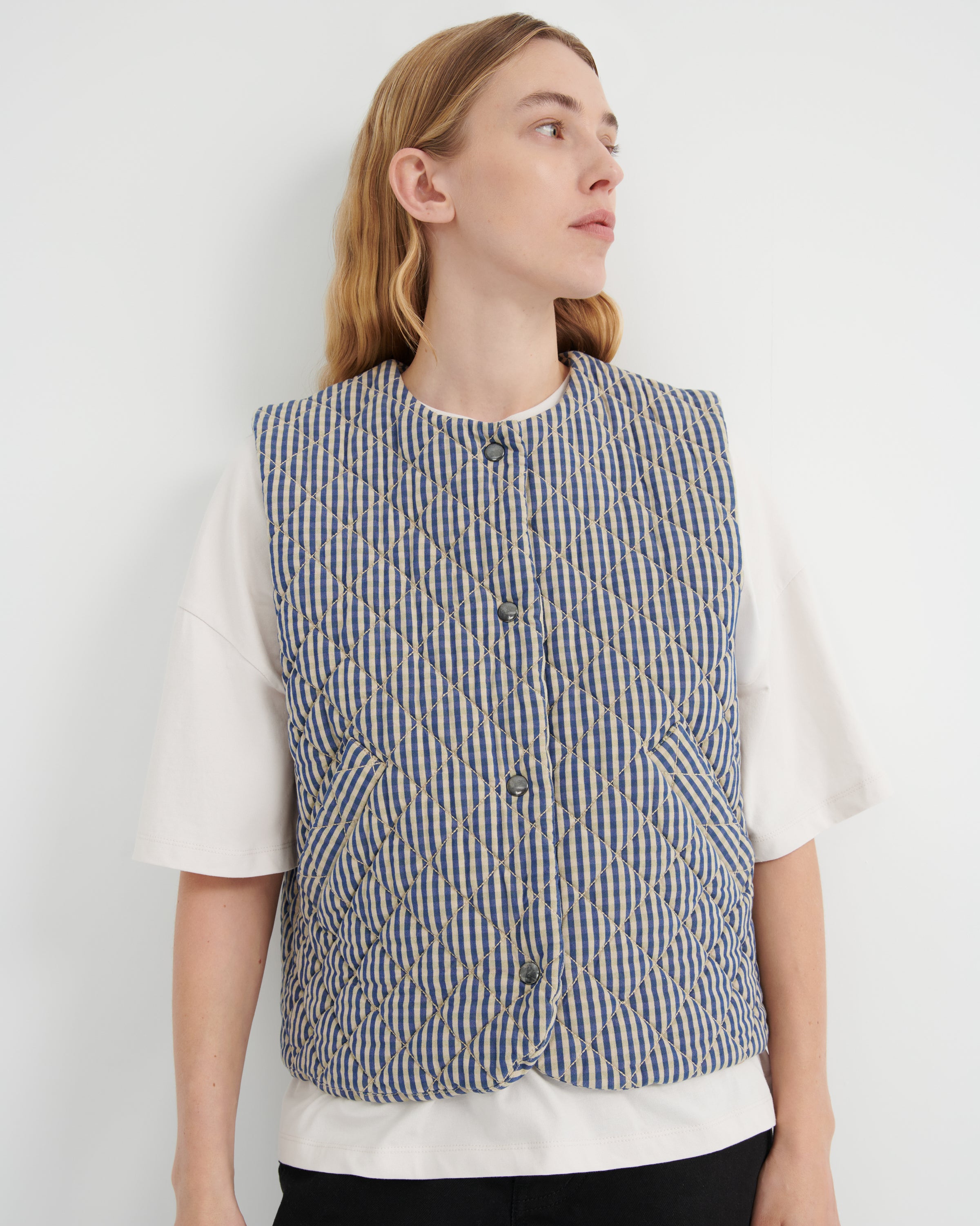 A woman wearing a sleeveless quilted vest with vertical blue, beige, and white stripes over a white short-sleeve top, photographed against a plain light gray wall.