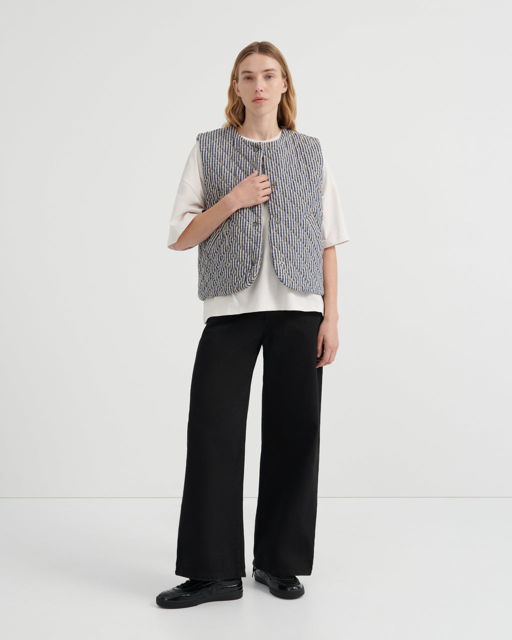 A model stands against a plain white studio background wearing a sleeveless textured gray vest over a white short-sleeve top, black wide-leg trousers, and black shoes.