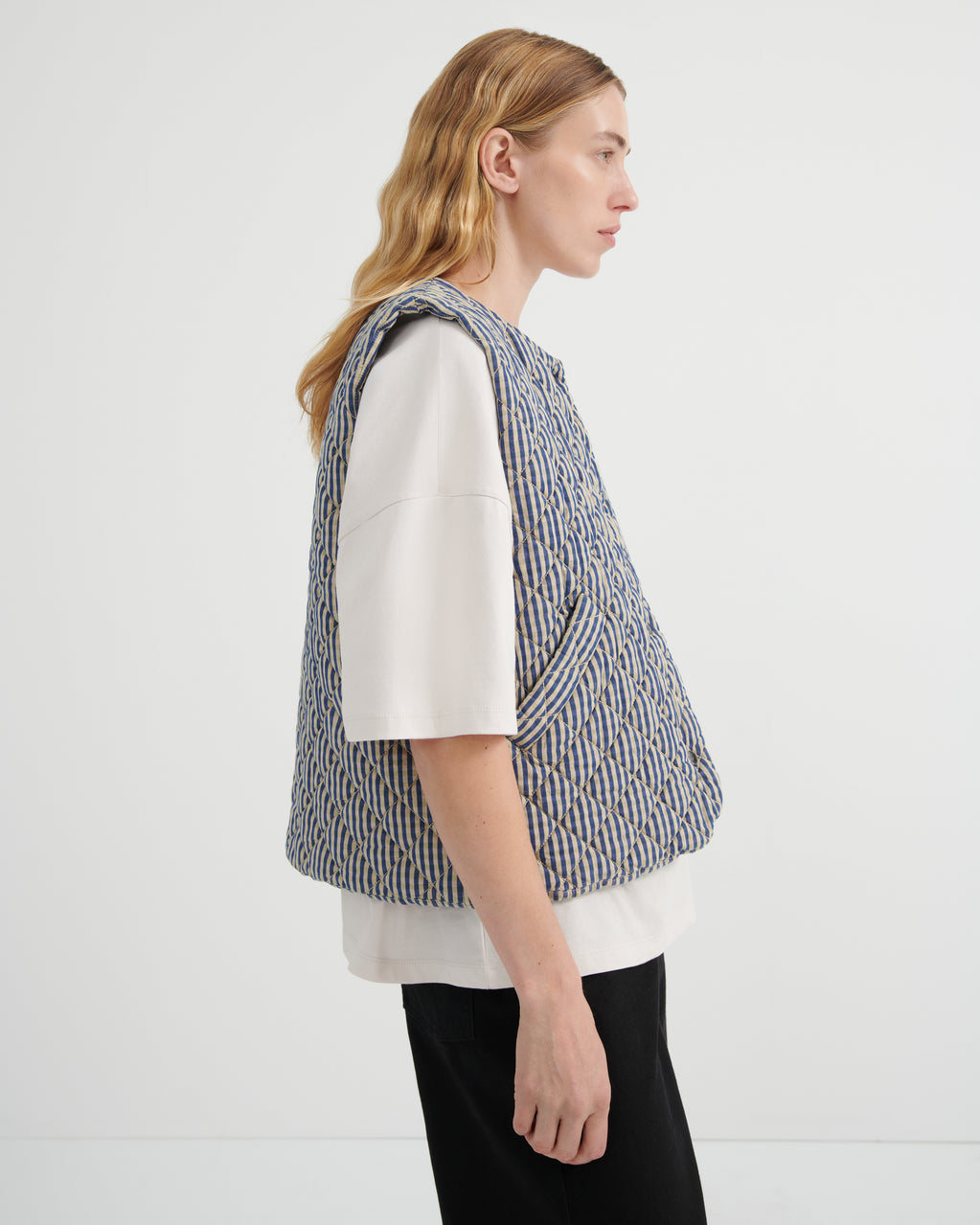 A model shown from the side profile wearing a blue and white vertically striped quilted vest over a white short-sleeve top and black pants, set against a plain white studio background.