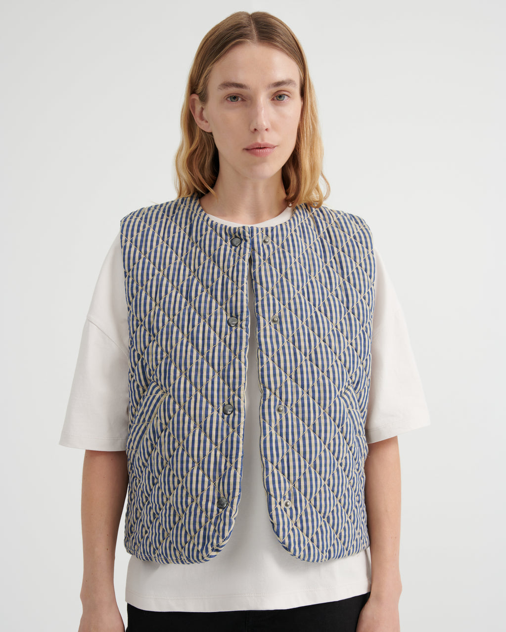 A model wearing a blue and beige vertically striped quilted vest over a white t-shirt, photographed against a light gray studio background.
