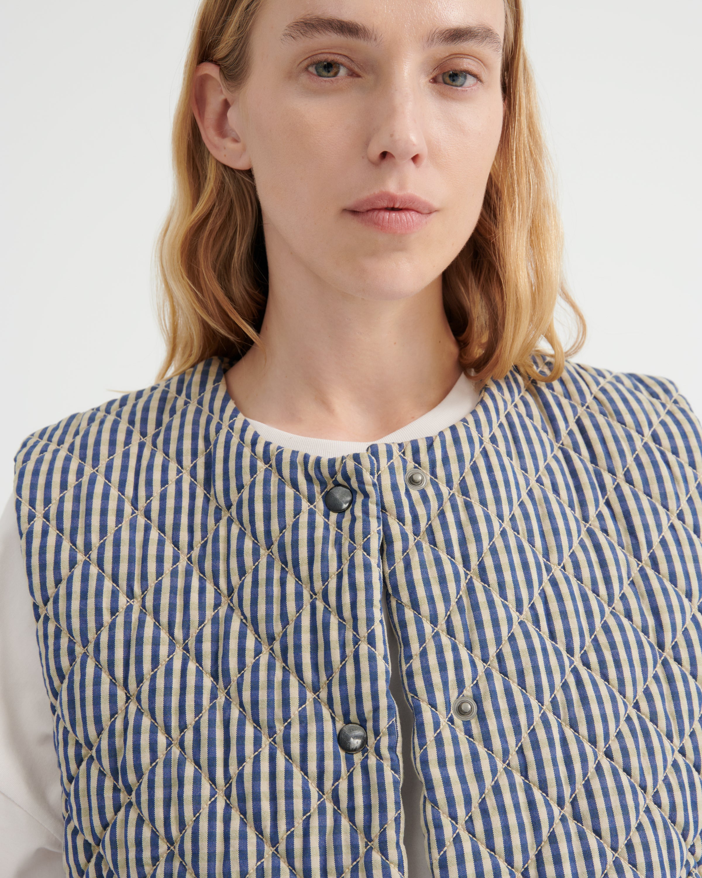 A close-up of a model wearing a blue and white gingham quilted vest with a cream-colored diamond-stitched pattern over a white crewneck shirt.