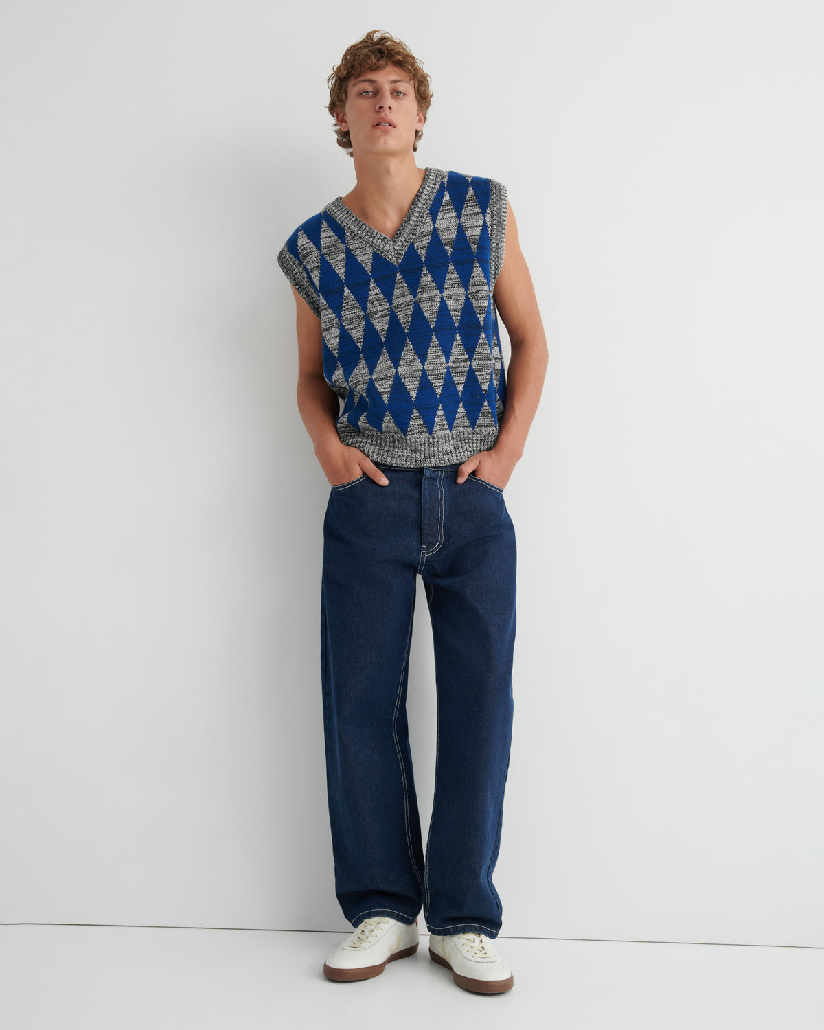 A model wearing a sleeveless knit sweater vest with a blue and gray geometric diamond pattern, dark blue jeans, and white sneakers.