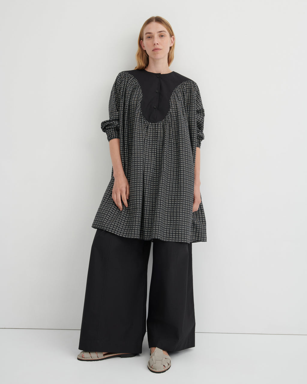A woman modeling a black and white micro-check tunic top with a solid black yoke and button placket, paired with wide-leg black trousers and beige flat sandals, against a plain white studio background.