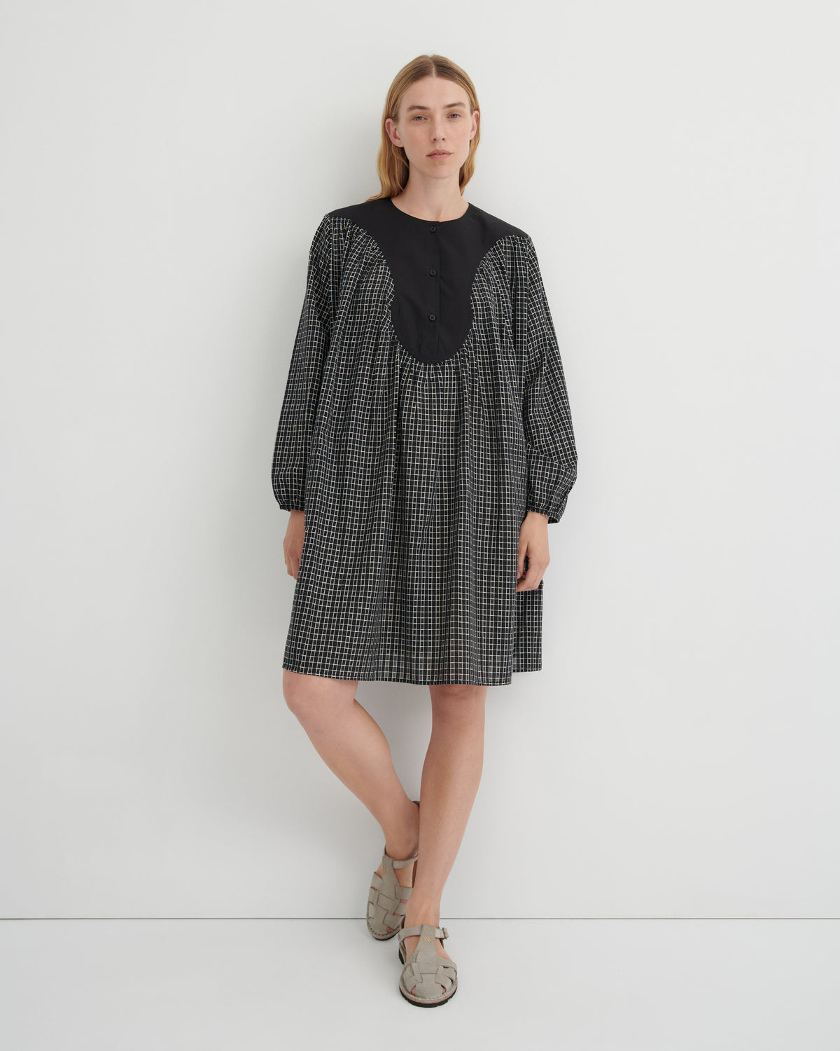 A woman modeling a black and white micro-check dress with a solid black round neckline and buttoned front panel, paired with beige flat sandals, against a plain white studio background.