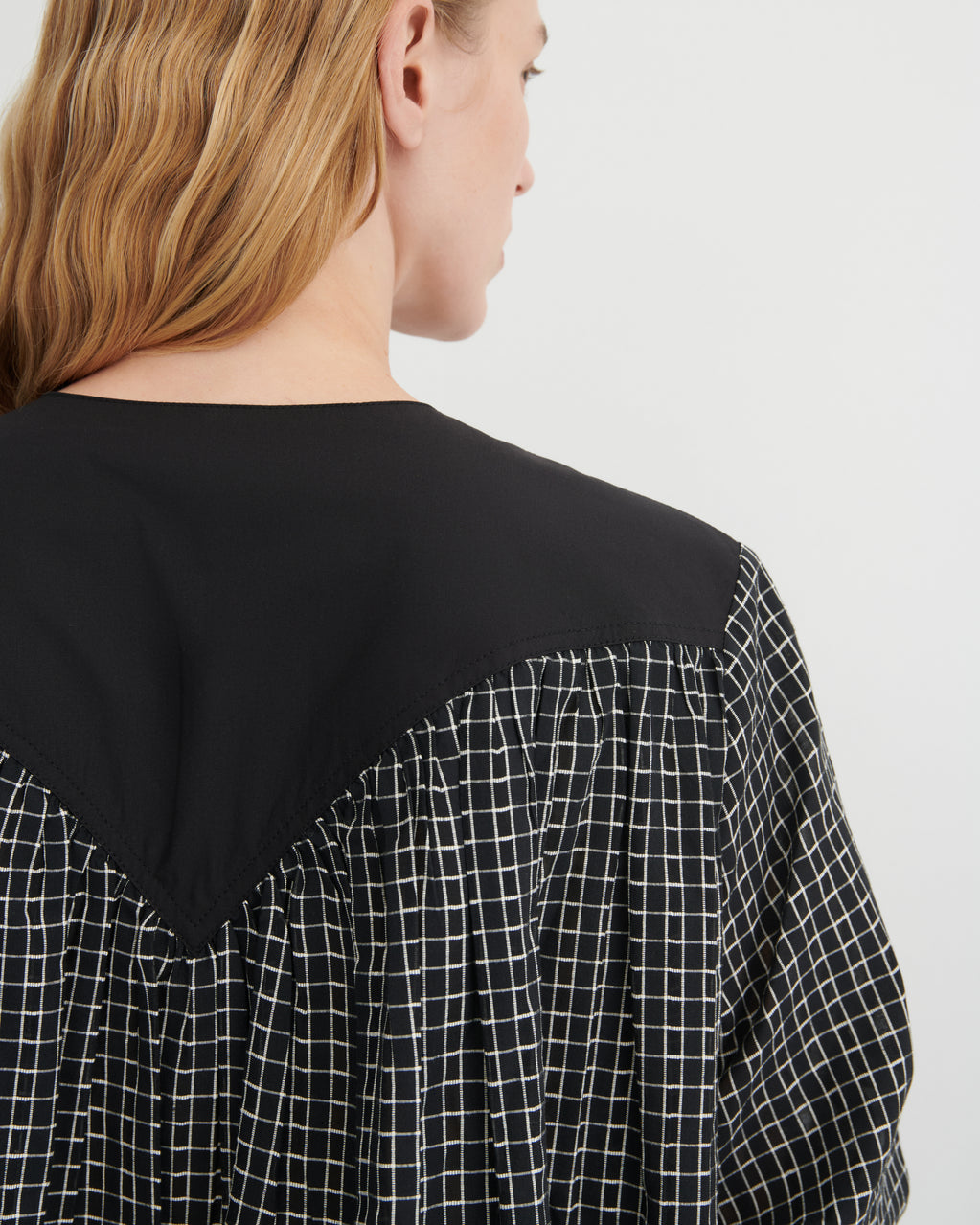 Close-up back view of a woman wearing a black blouse with a contrasting black-and-white windowpane check patterned lower back and sleeves, against a plain light gray background.
