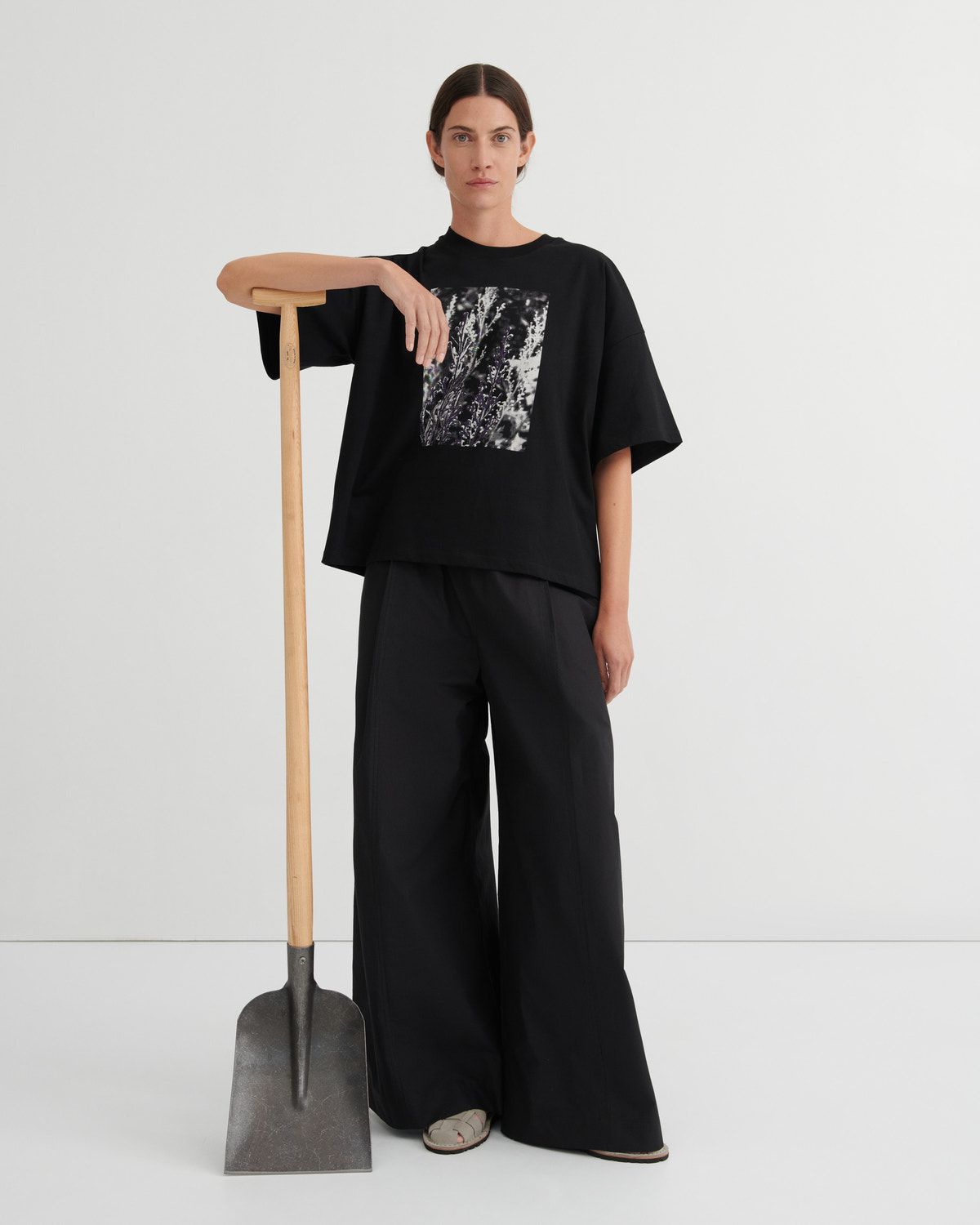 Person wearing a black outfit with a graphic design, standing next to a shovel on a white background