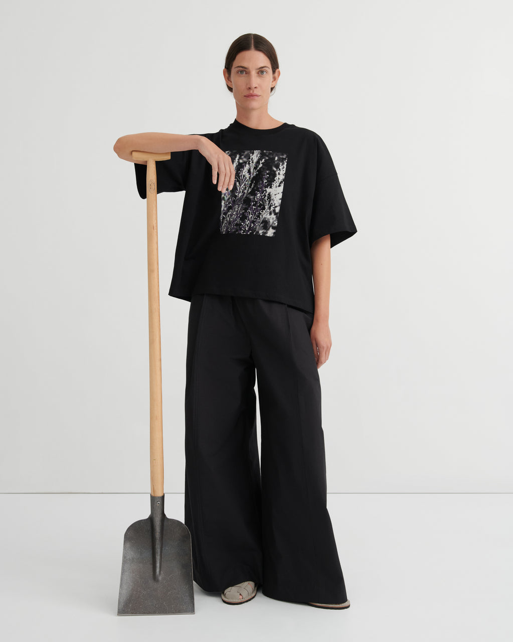 Person wearing a black outfit with a graphic design, standing next to a shovel on a white background