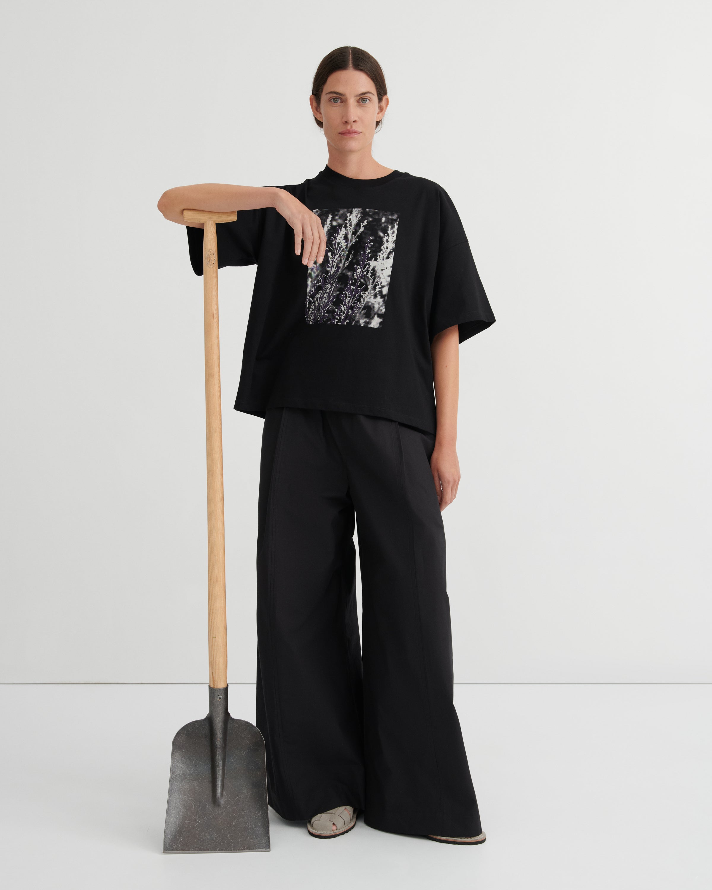 Person wearing a black outfit with a graphic design, standing next to a shovel on a white background