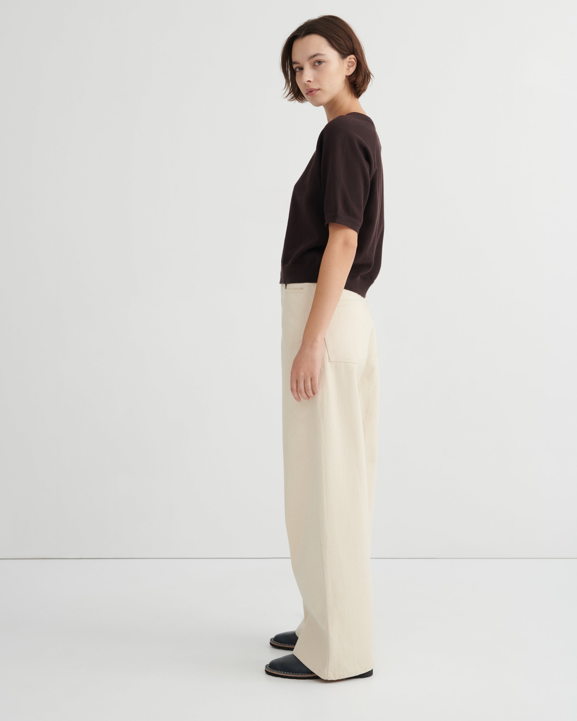 A woman stands in profile wearing a dark brown short-sleeve top, cream wide-leg trousers, and dark loafers against a clean white studio background.