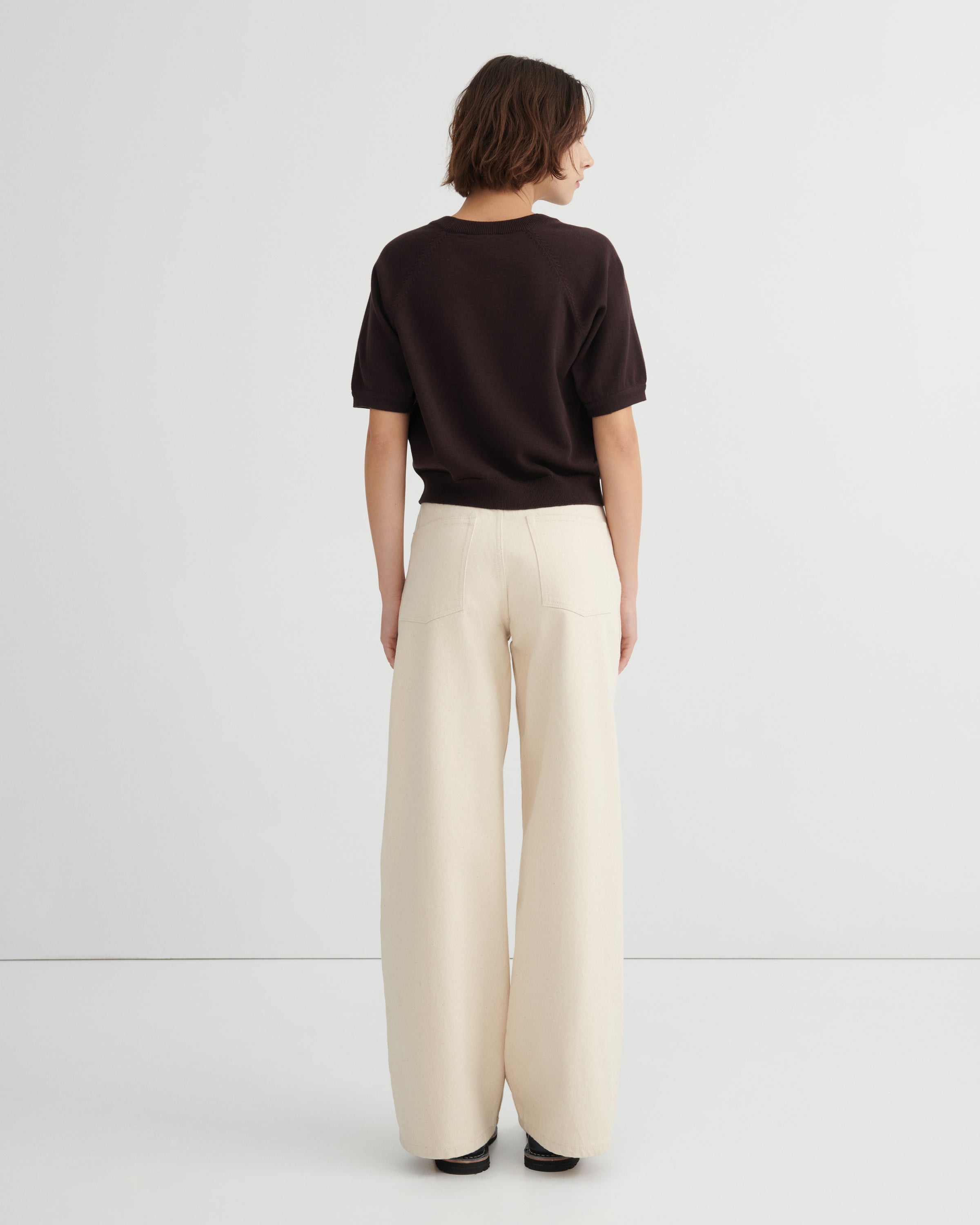 A model shown from the back wearing a dark brown short-sleeve top and cream wide-leg trousers against a light gray studio background.