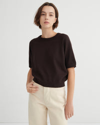 A model wearing a dark brown short-sleeve crewneck sweater and cream-colored trousers, standing against a light gray studio background.
