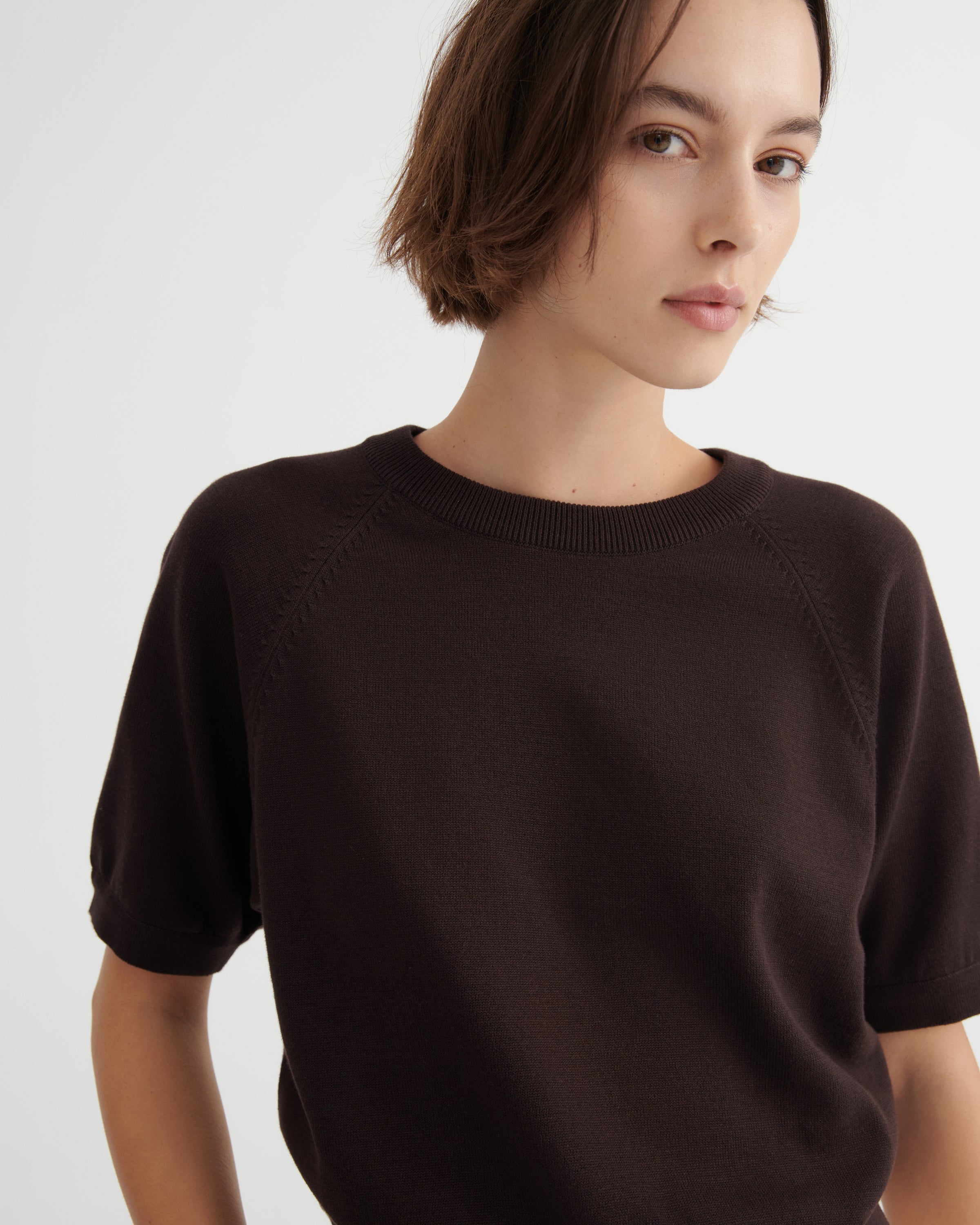 A close-up of a model wearing a dark brown short-sleeve knit sweater with raglan sleeves against a light gray background.