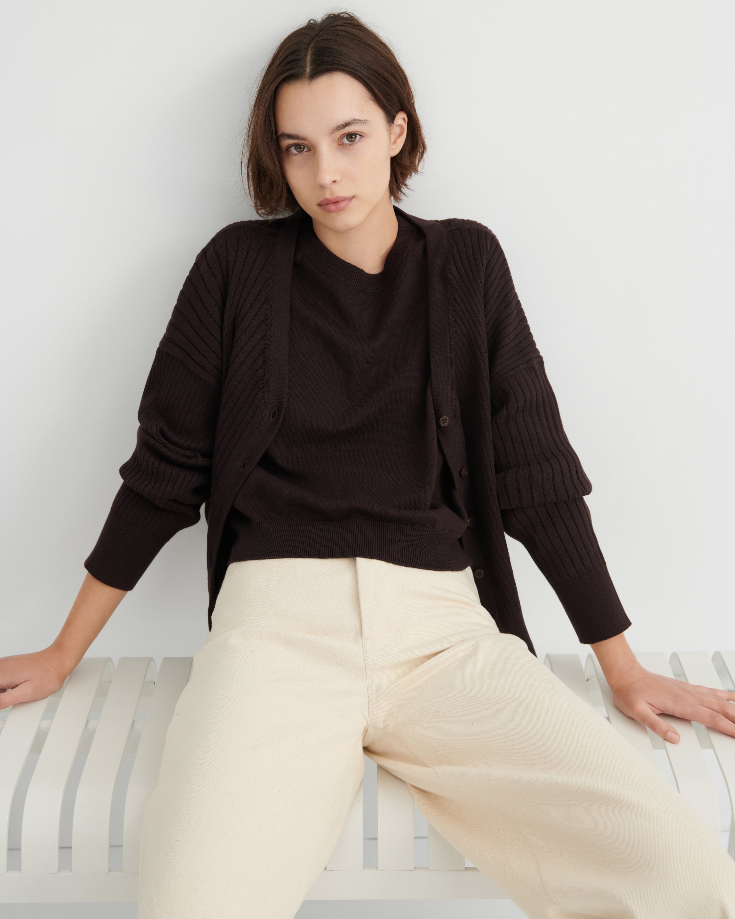 A woman wearing a dark brown ribbed cardigan over a matching dark brown top and cream wide-leg trousers, sitting on a white slatted bench against a light gray wall.