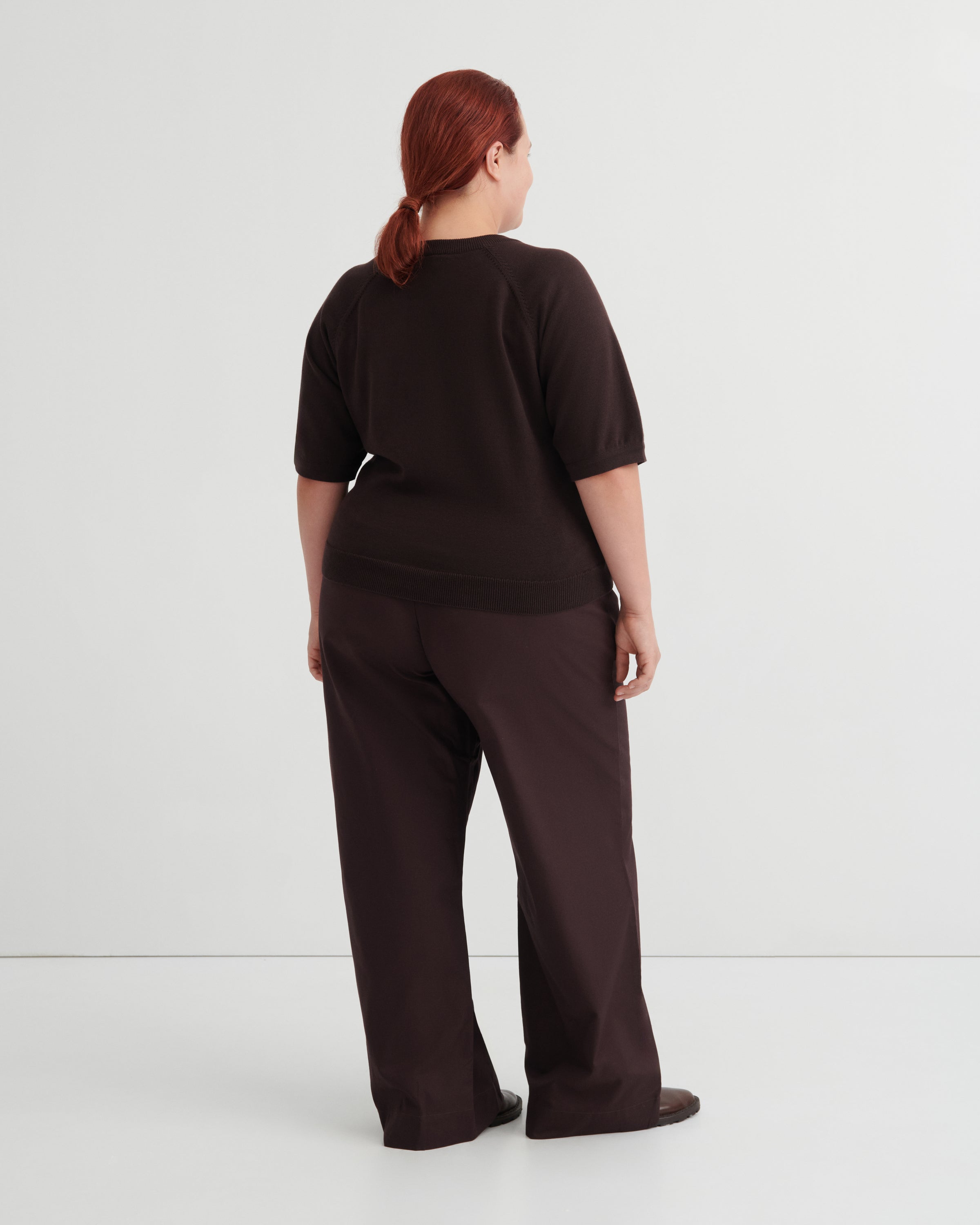 A model with red hair tied back is shown from behind wearing a dark brown short-sleeve top and matching wide-leg trousers against a light gray studio background.