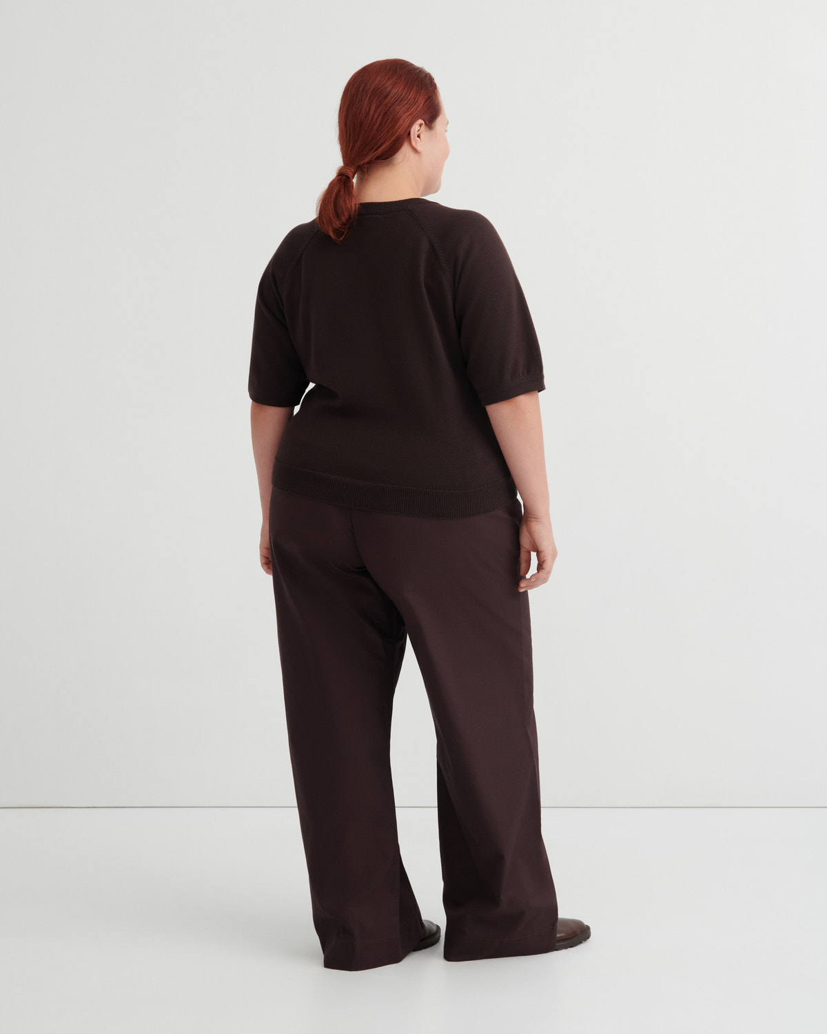 A model with red hair tied back is shown from behind wearing a dark brown short-sleeve top and matching wide-leg trousers against a light gray studio background.